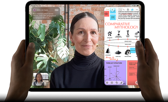 iPad Pro, front exterior, landscape orientation, space black color, rounded corners, black display bezel, hands holding left and right sides, display showing a FaceTime conversation being live translated, and an educational app showing a presentation of "Comparative Mythology" with text, diagrams, and handwritten notes