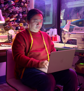 A person uses MacBook Neo, blush color, unplugged on their lap, in a workspace with a sewing machine and related materials, including thread and a dress form
